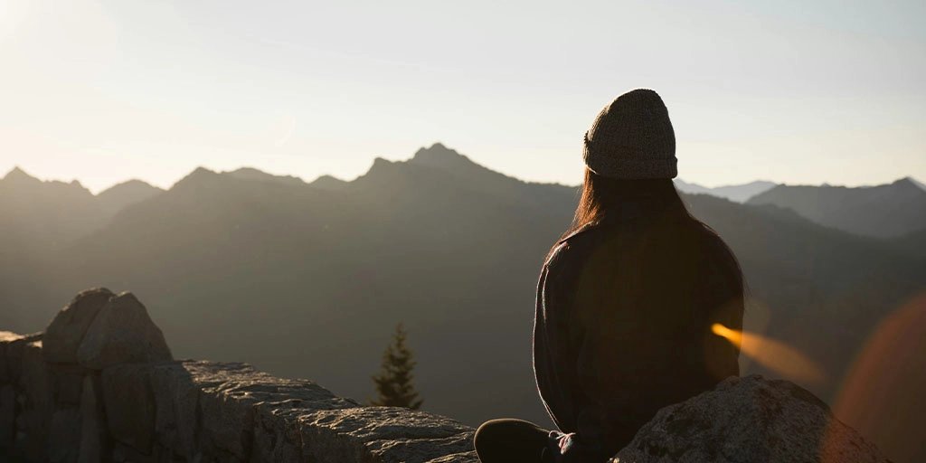 Backlit hiker in a beanie sits on a rocky mountain ridge, facing jagged peaks at sunrise; golden lens flare creates a quiet, contemplative mood.