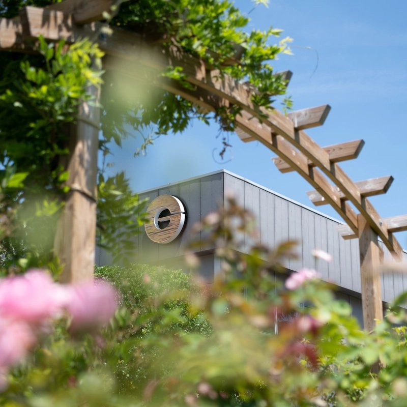 Sunlit garden with pink flowers and leafy vines frames the Givaudan Argentuil Perfumery School. Wooden pergola, and round window beneath a blue sky.