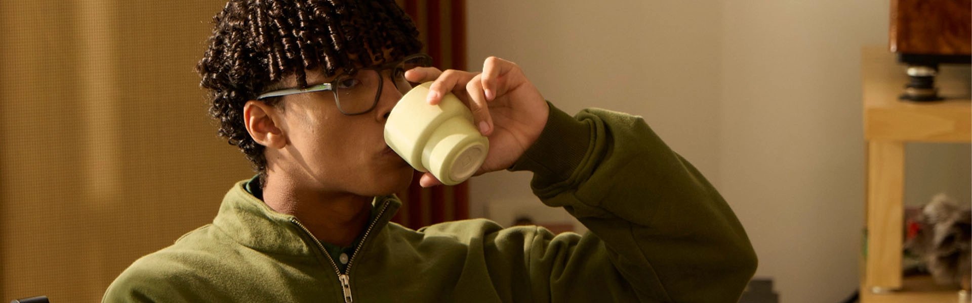 A relaxed person in an olive-green sweater and jeans sits cross-legged in a sunlit room, sipping from a cream mug; warm light creates a calm, quiet mood.