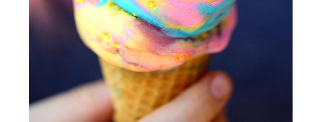 Bright, swirled ice cream in vibrant shades of blue, pink, and yellow is close-up, showcasing its creamy texture. The background is softly blurred, highlighting the indulgent treat.