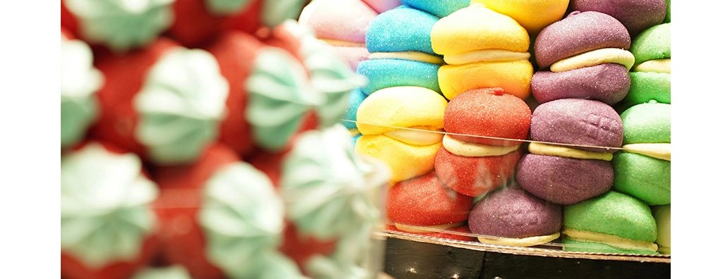 A close-up view of a colourful striped cake showcasing layers of purple, turquoise, green, yellow, orange, and red sponge, with a light frosting visible between the layers. The vibrant colours create a joyful, festive appearance.