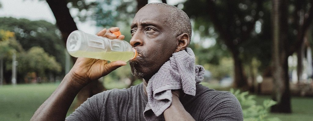 A man drinks from a clear water bottle in a green park, towel around his neck, looking tired and focused after an outdoor workout.