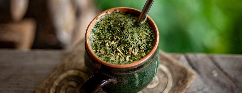 A rustic green ceramic mate cup, filled with yerba leaves and a metal straw, rests on a weathered wooden table amid softly blurred greenery.