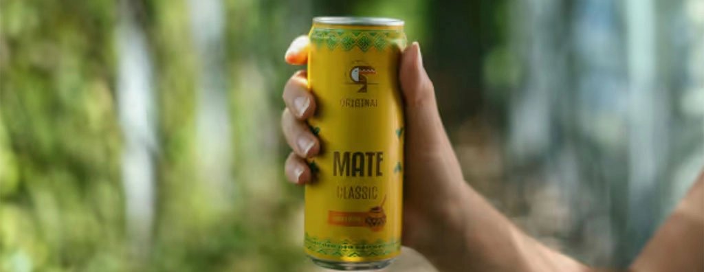 A hand holds a yellow can of Yerba Mate Classic outdoors, sharply lit against a softly blurred green garden background, creating a fresh, relaxed mood.