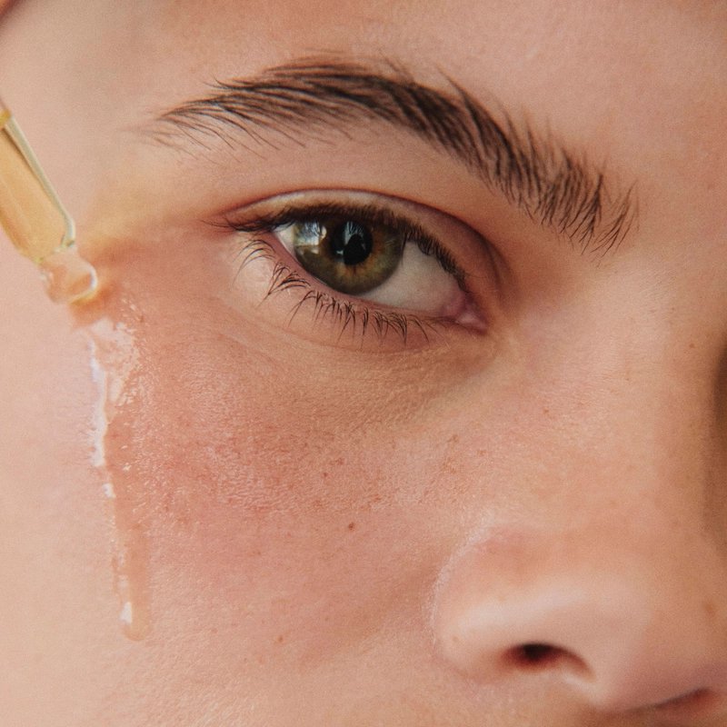 Close-up of a green-brown eye as a glass dropper applies skincare serum to the cheek; dewy skin and soft lighting convey a calm beauty routine.