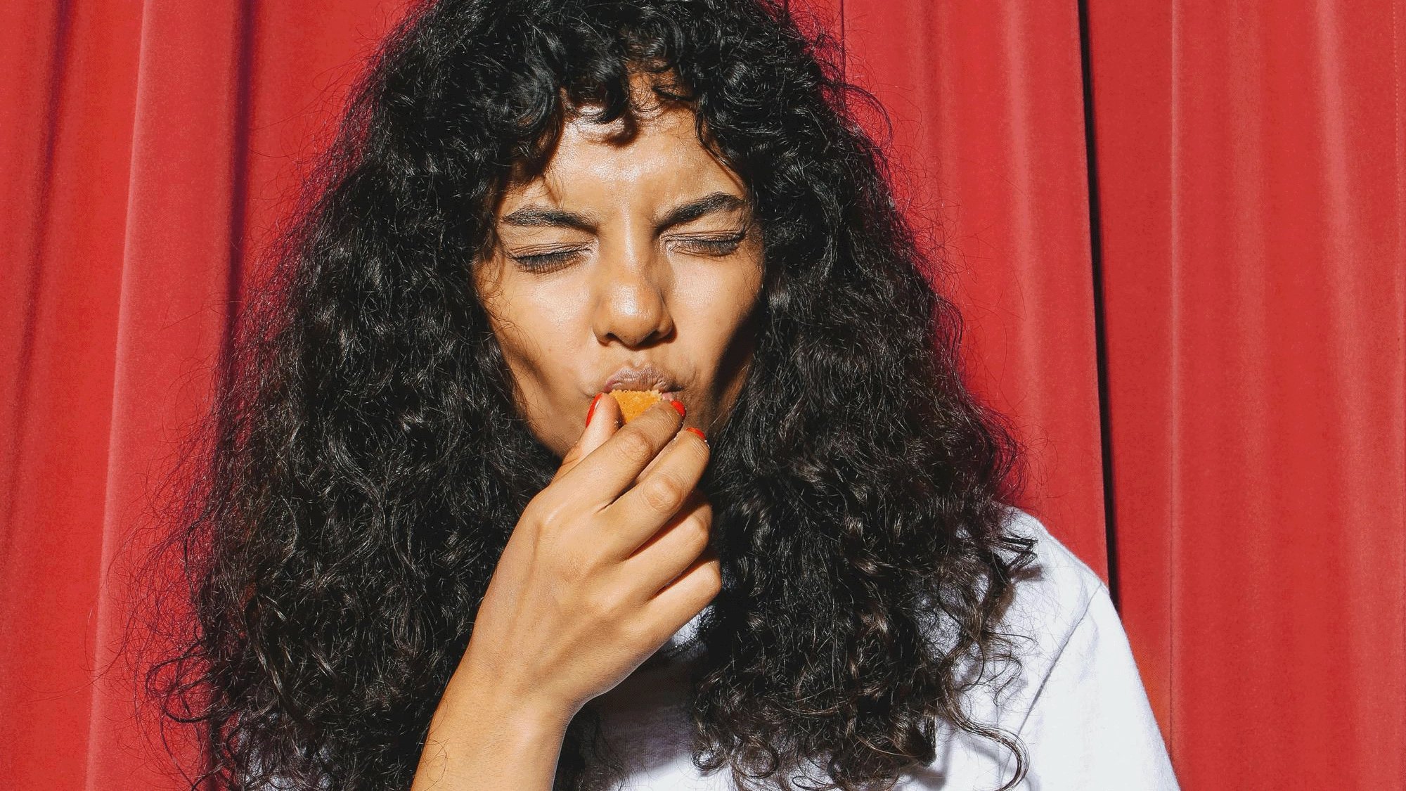 A woman with long, curly dark hair closes her eyes while biting a small red snack, framed by vivid red curtains in a quiet, intimate moment.