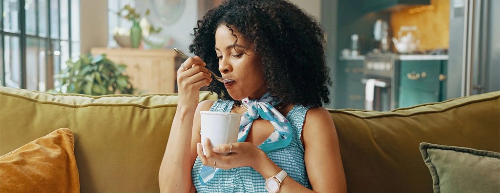 A relaxed woman sits on a mustard-yellow sofa, sipping a drink from a cup in a bright home interior, conveying a calm, cozy moment.