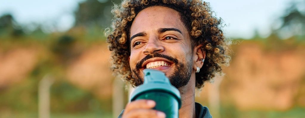 A smiling man with curly hair and a beard holds a teal water bottle outdoors, enjoying a cheerful moment against a softly blurred green landscape.