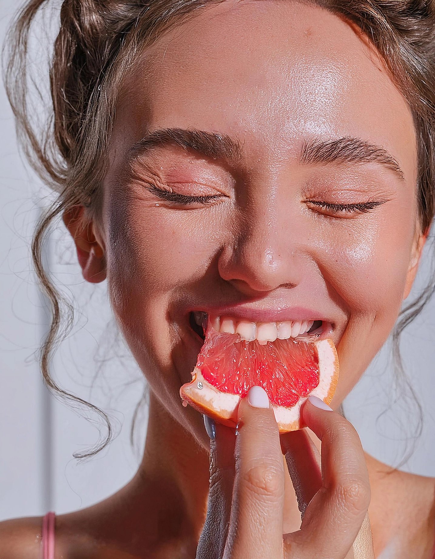 Close-up of a smiling woman with closed eyes biting a juicy red grapefruit wedge, sunlight highlighting her face and fresh, playful mood.
