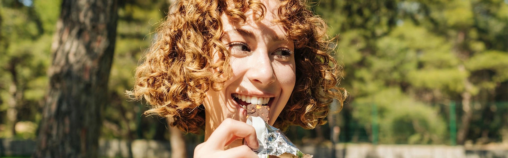 A young curly-haired woman beams as she bites a chocolate bar in silver foil, enjoying a carefree sunny moment among green trees.