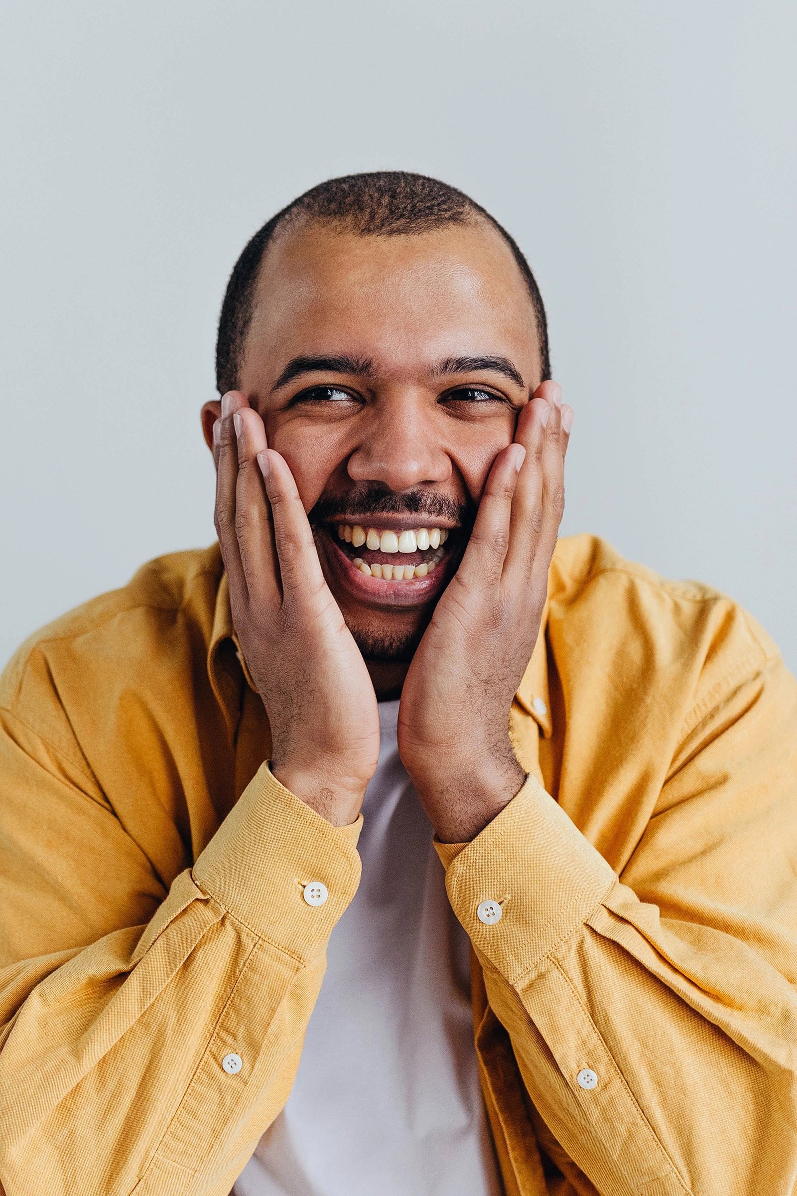 A smiling man in a mustard-yellow shirt presses both hands to his cheeks against a plain gray background, conveying joyful surprise and excitement.