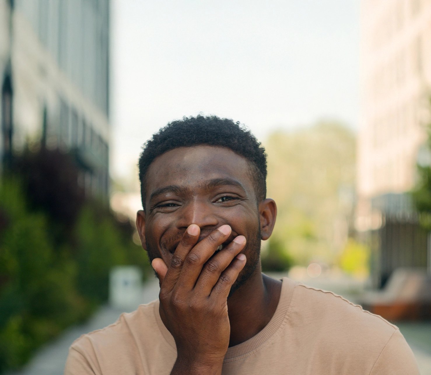 A young man in a beige shirt covers his mouth with one hand, smiling with amused surprise on a sunlit city street; the mood is warm and playful.