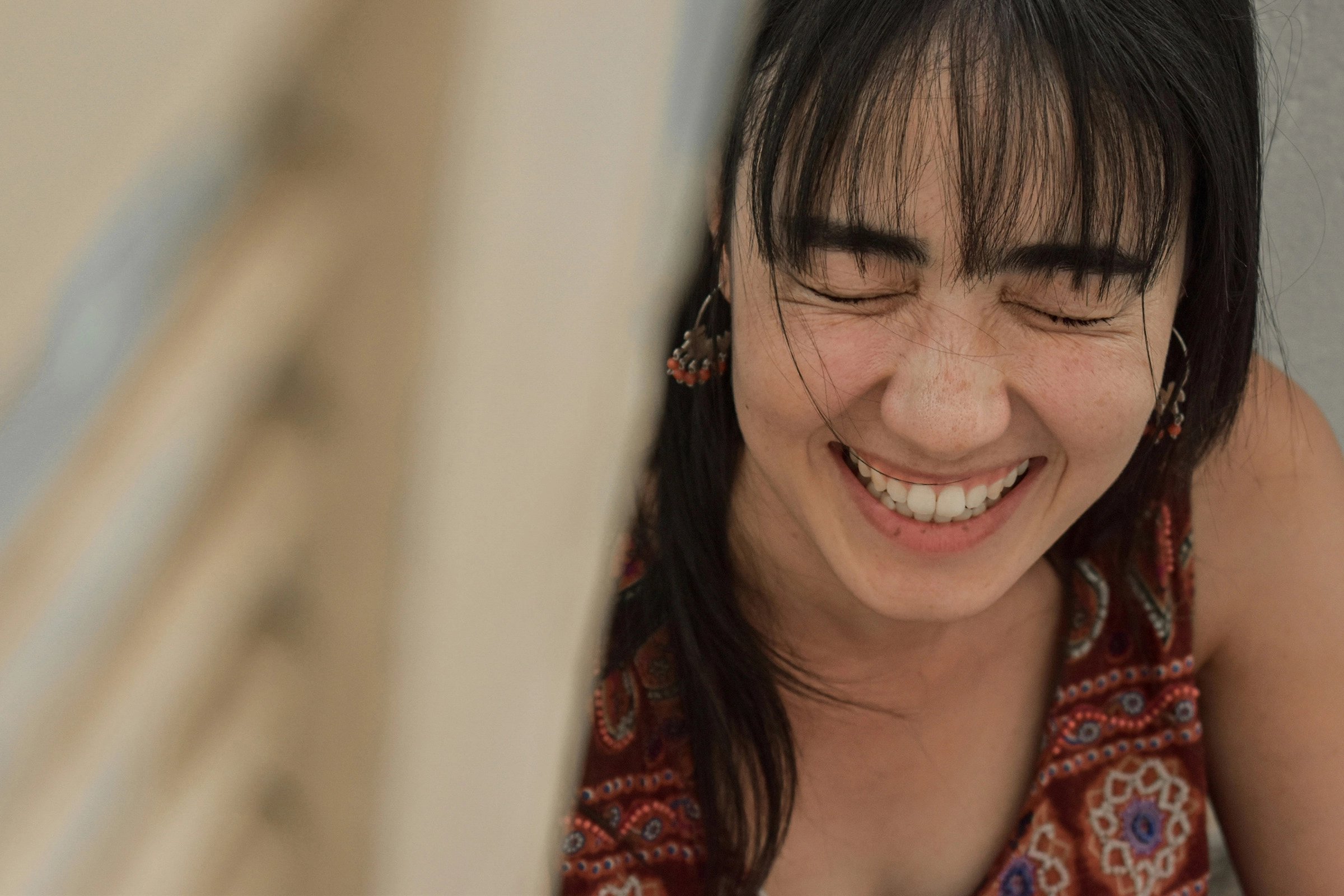 A young woman in a patterned red top laughs with her eyes closed, her bangs falling across her face; the close-up feels joyful and candid.