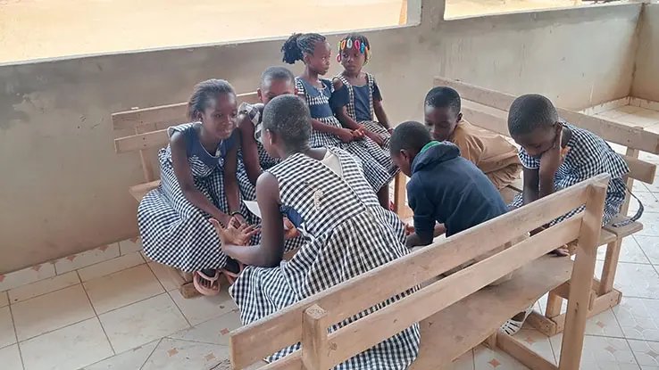 A group of children in checkered dresses are seated on wooden benches in a classroom. They are engaged in a discussion, with som